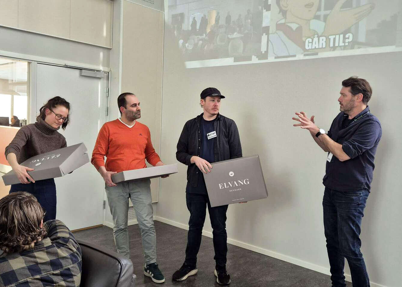 Tre personer holder hver sin eske som det står "Elvang" på, og en fjerde person snakker til dem mens han gestikulerer.