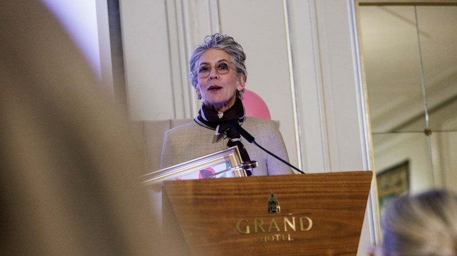Kvinne på talestol merket Grand Hotel, holder tale, holder et diplom
