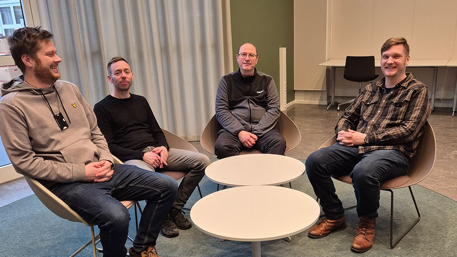 Foto av fire menn som sitter rundt to lave, runde bord i et hyggelig rom. Mennene smiler og ser blide ut. 